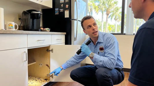 An exterminator wearing blue gloves and a uniform inspects a Florida commercial office break room, using a flashlight and pointing to evidence of a pest problem inside an open lower cabinet.