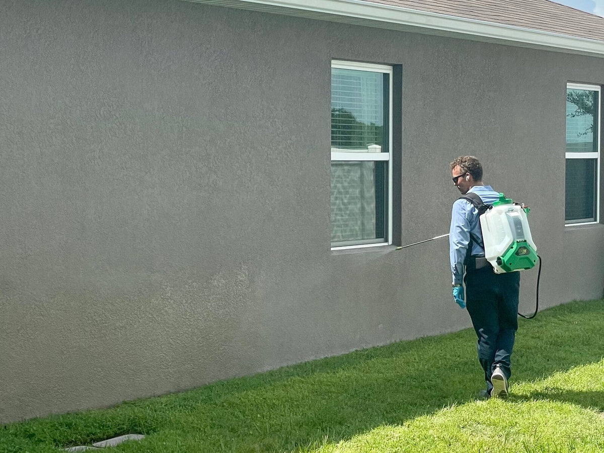 A Webenex technician spraying the foundation and windows of a gray stucco home with a backpack sprayer